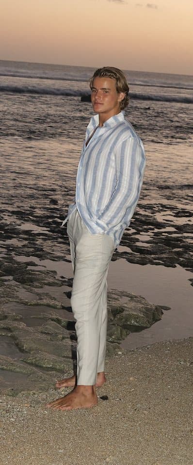 The coach standing barefoot on the shoreline at sunset in a striped linen shirt and white trousers